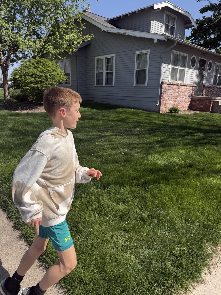 A boy in a tan hoodie and teal shorts runs past a gray house on a bright, sunny day.