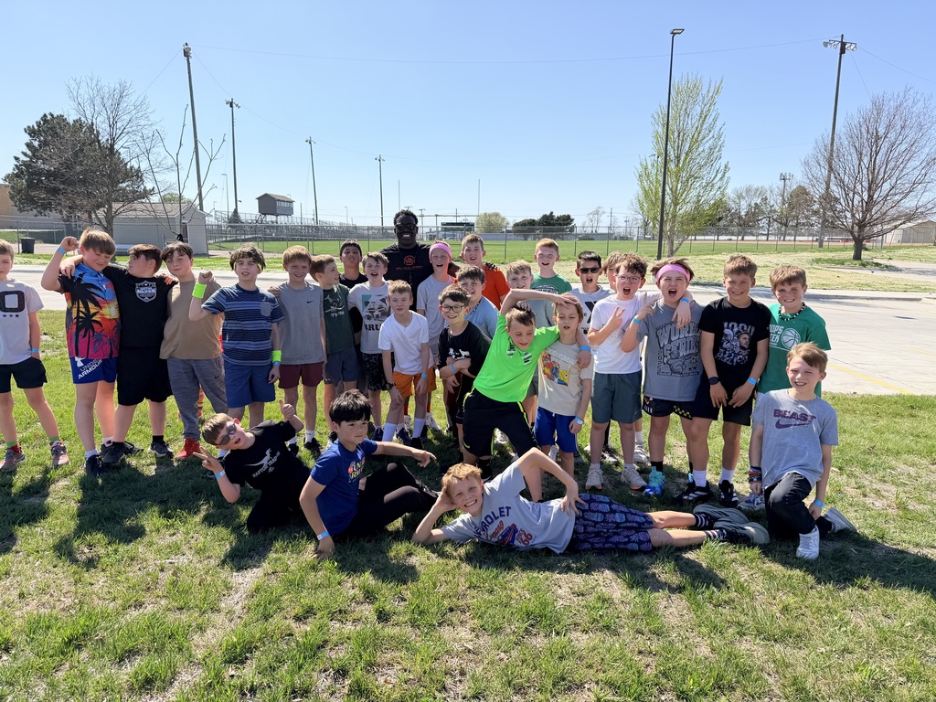 A large group of elementary-aged boys and a man pose for a group photo on a grassy field under a clear blue sky.