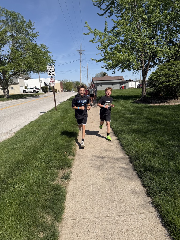 Two boys in athletic gear run toward the camera on a neighborhood sidewalk lined with green grass and trees.