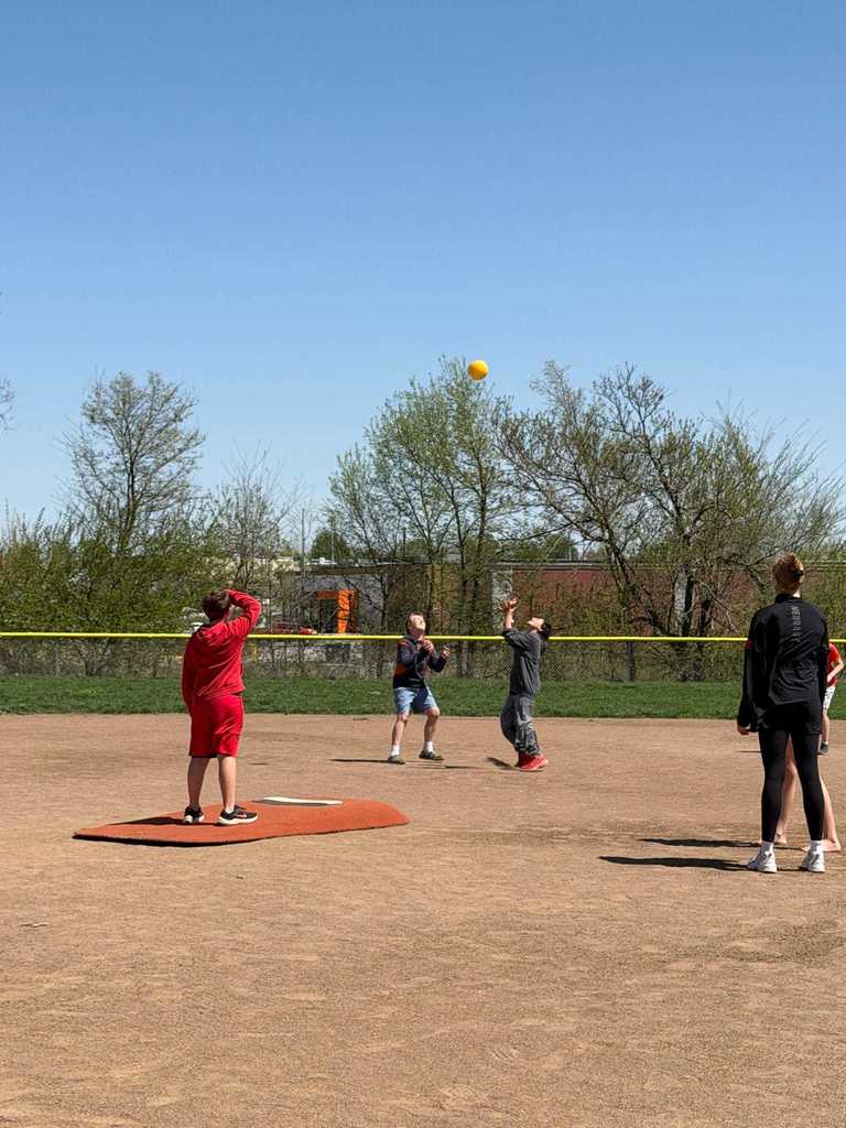 Two children looking up at a high yellow ball in the air on a dirt baseball field.