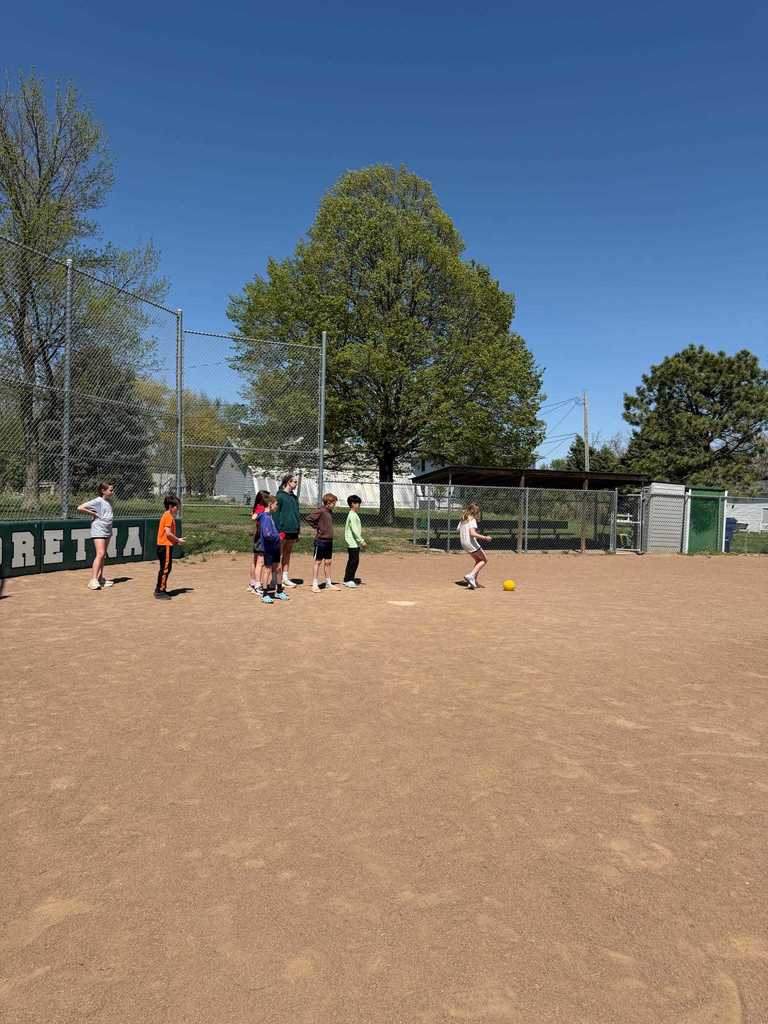 A girl kicking a yellow ball toward a line of children waiting their turn on a dirt field.
