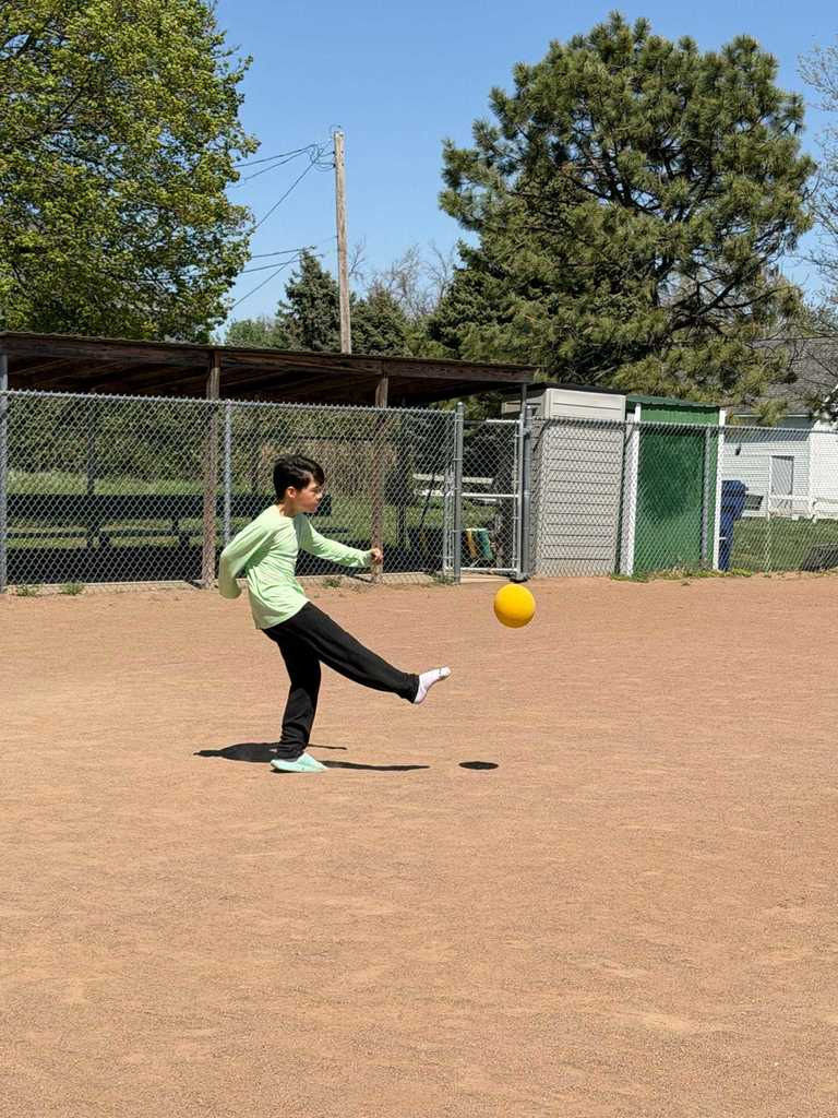 A boy in a light green long-sleeve shirt kicking a yellow ball on a sunny day.