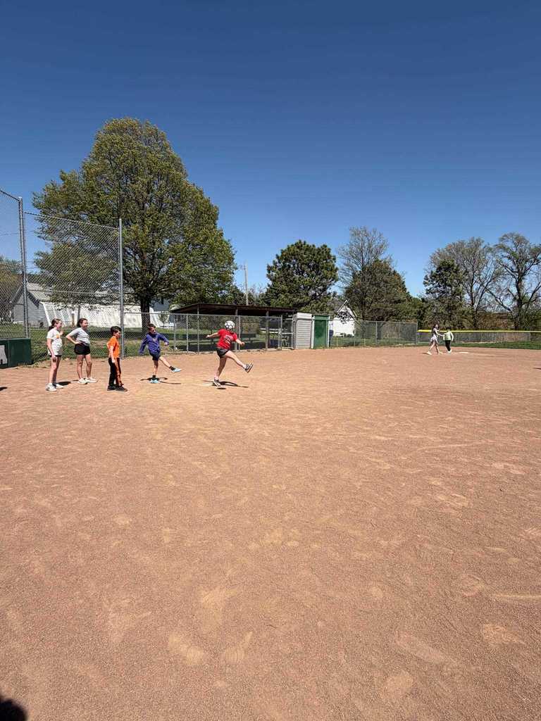 A girl in a red shirt kicking a ball on a wide dirt field with a large tree in the background.