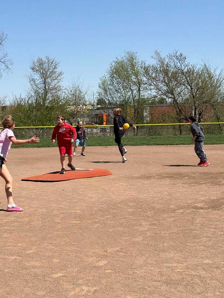 An adult in black chasing a yellow ball across the dirt as other kids look on.
