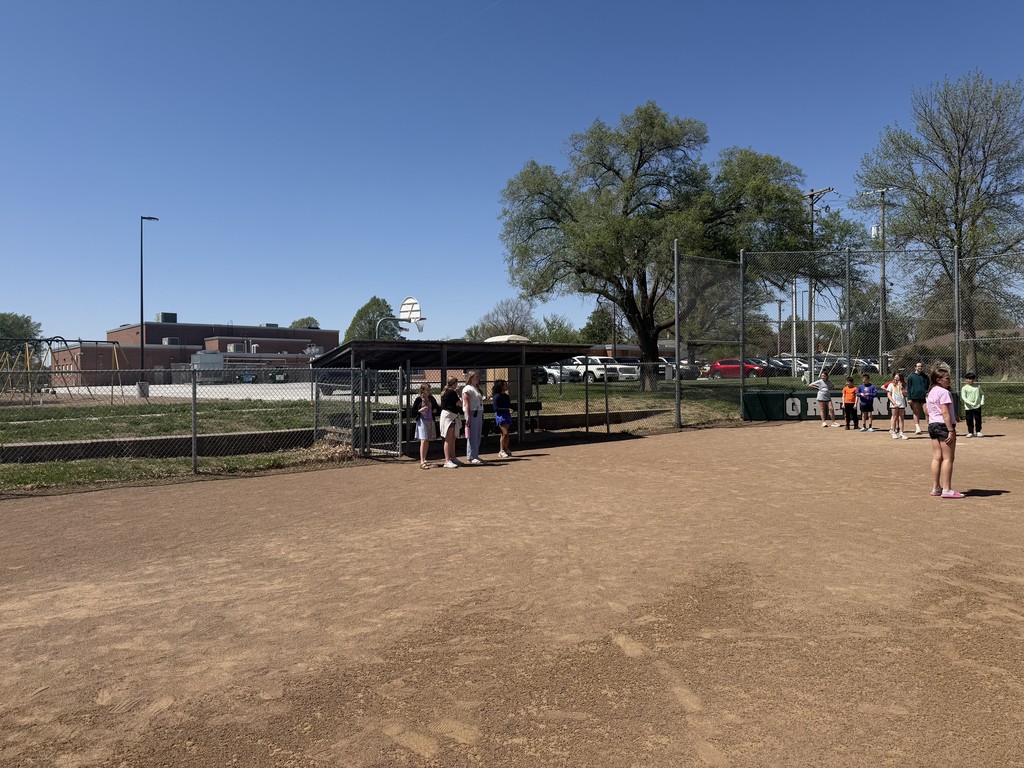 Several children standing near the dugout and home plate area of a dirt baseball field.
