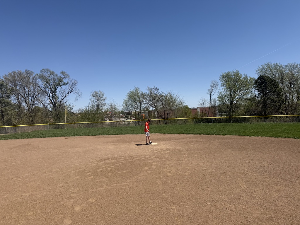 A single child in a red shirt standing on a base in the middle of a vast dirt field.