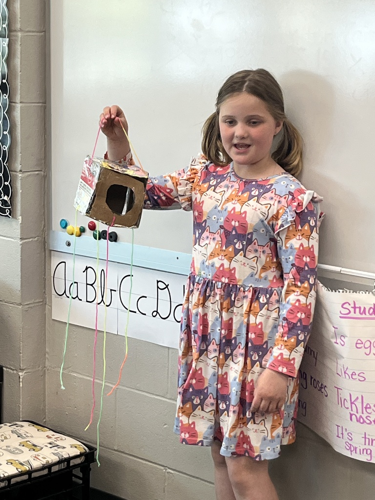 A girl in a cat-patterned dress holds up a cardboard birdhouse project decorated with colorful hanging strings.