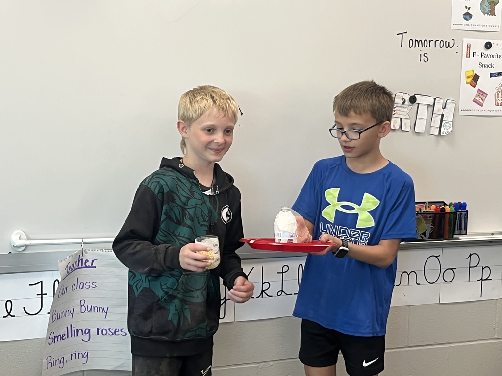 Two boys present a project featuring a plastic bottle wrapped in white material sitting on a red plate.