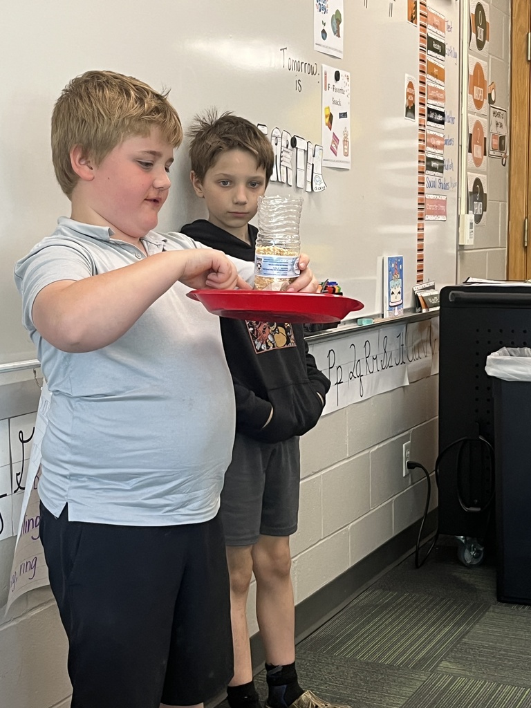 Two boys stand by a whiteboard; one holds a red plate supporting a clear plastic bottle filled with birdseed.
