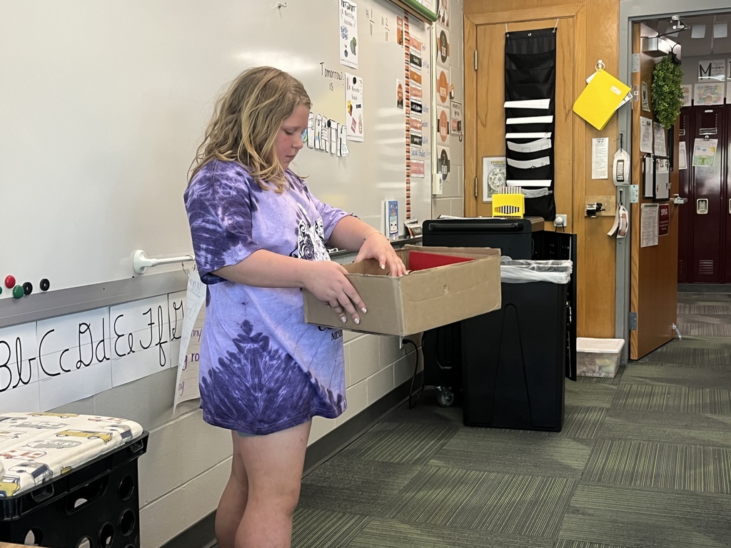 A girl in a purple tie-dye shirt stands in a classroom holding a large, shallow cardboard box.