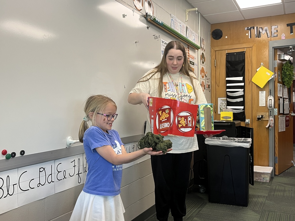 A teacher and a young girl stand in a classroom; the teacher holds a Cheez-It box project while the girl holds a bundle of green yarn.