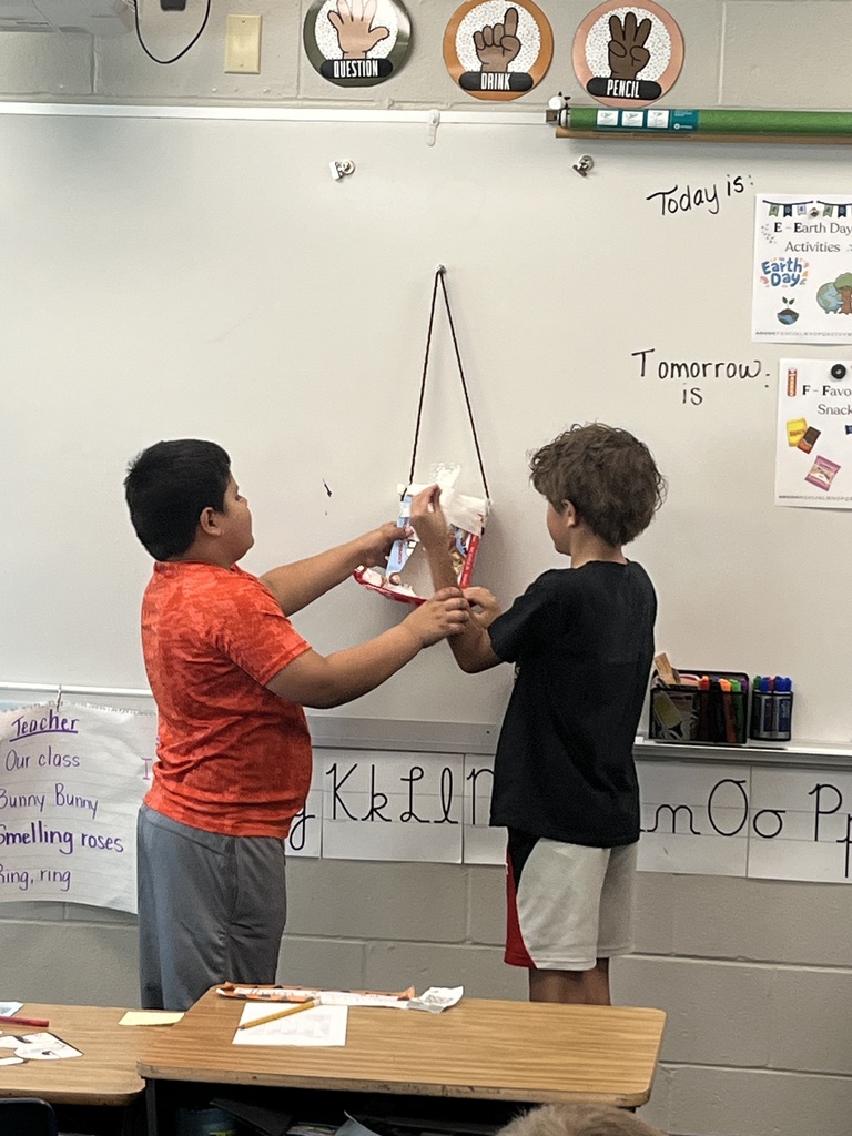 Two boys stand at a whiteboard working with a hanging recycled-material project made from a snack box and string.
