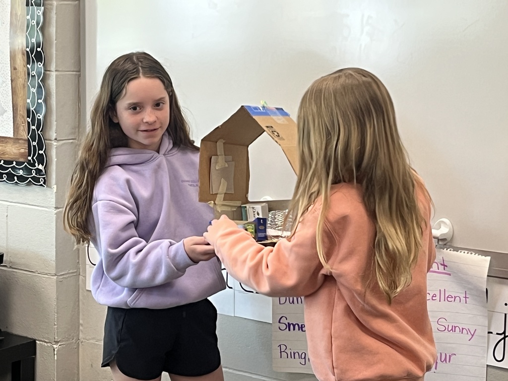Two girls examine a handmade cardboard structure that resembles a small house or diorama.