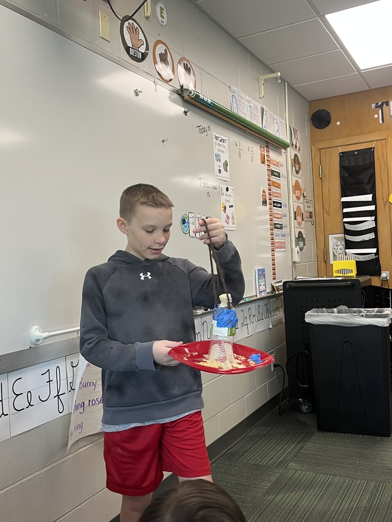A young boy in a grey hoodie holds a red plate with a bird feeder made from a plastic bottle and string in front of a classroom whiteboard.