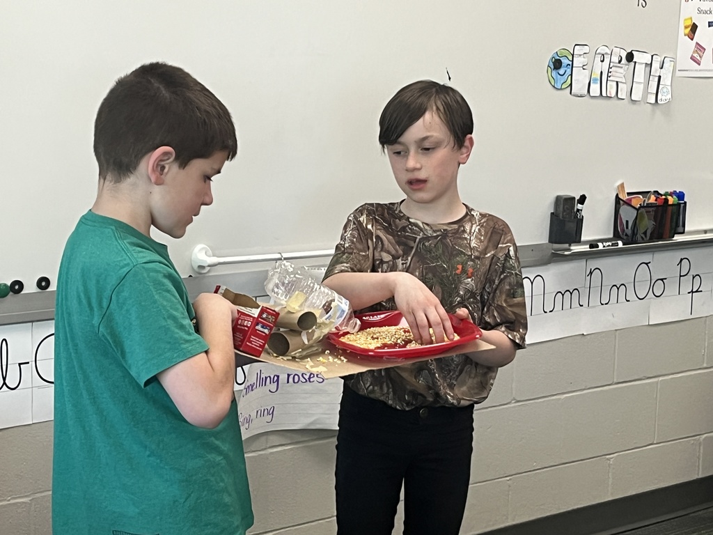 Two boys collaborate on a project involving a red plate, cardboard tubes, and a plastic bottle containing seeds.