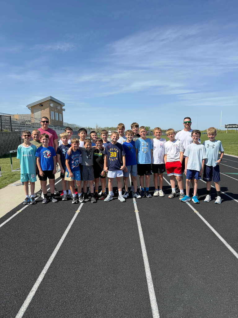 A group of about 20 young boys and two adult male coaches standing together on a black running track for a team photo. The boys are wearing various athletic shirts and shorts, with some wearing sunglasses. One boy in the center wears a navy "Golden State" jersey with the number 30. They are positioned in front of a stadium bleacher structure and a small press box. The sky is bright blue with light, wispy clouds, and the sun creates soft shadows on the track.