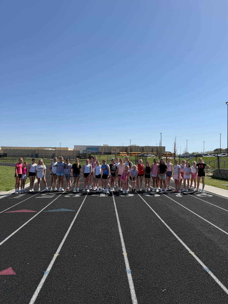 A wide-angle, eye-level shot of a large group of approximately 30 young girls standing in a long line across a black running track. They are dressed in colorful athletic wear—shorts, t-shirts, and tank tops—and are smiling at the camera under a bright, clear blue sky. In the background, a large school building and several yellow school buses are visible. The perspective of the white lane lines on the track converges toward the bottom of the frame.