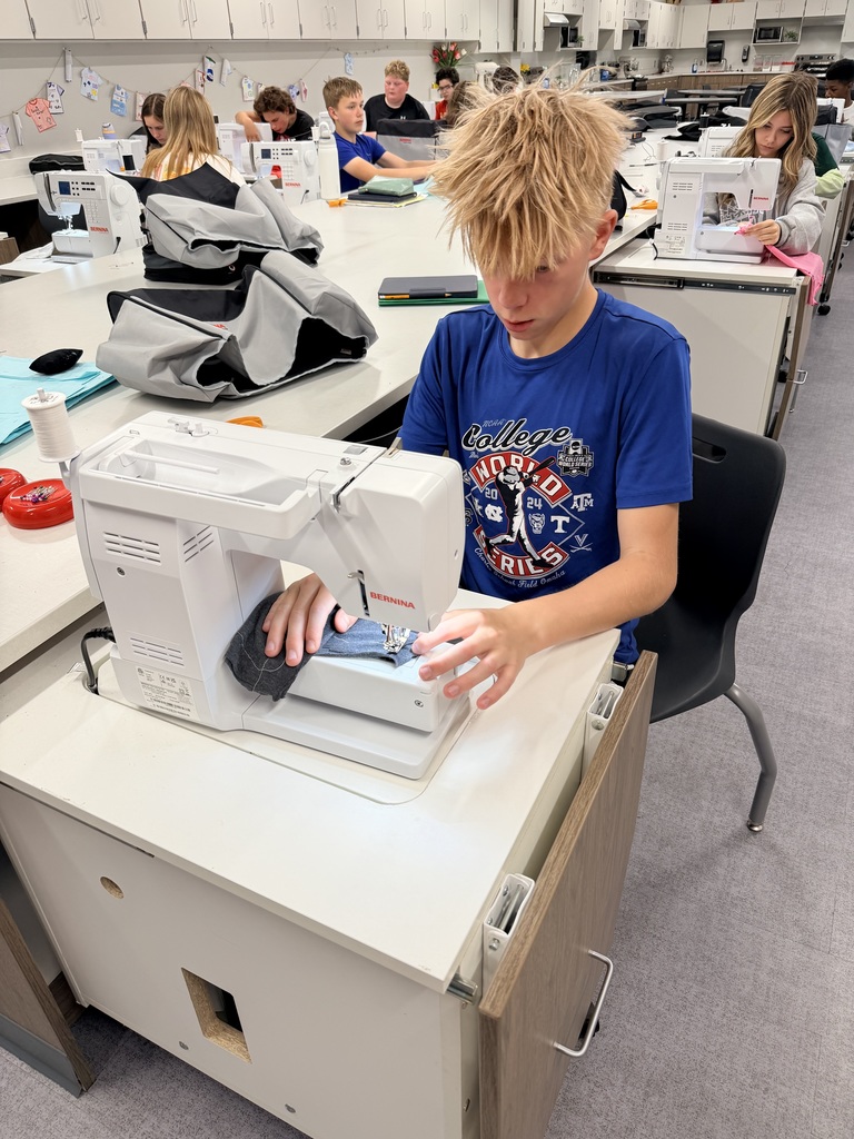 A male student sits at a sewing machine sewing. There are other students behind him sewing as well. 