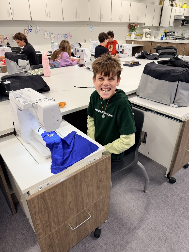 A male student sits at a sewing machine and smiles for the camera. 