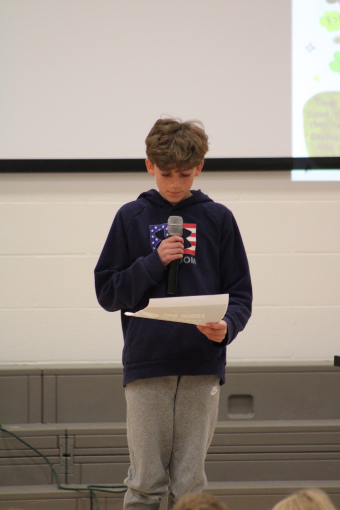 A boy in a navy blue hoodie stands at the front holding the microphone reading his poem.