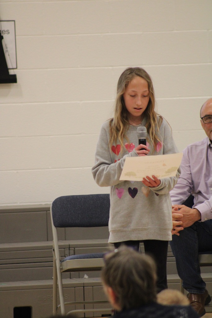 A girl stands at the front holding a microphone reading her Arbor Day poem. 