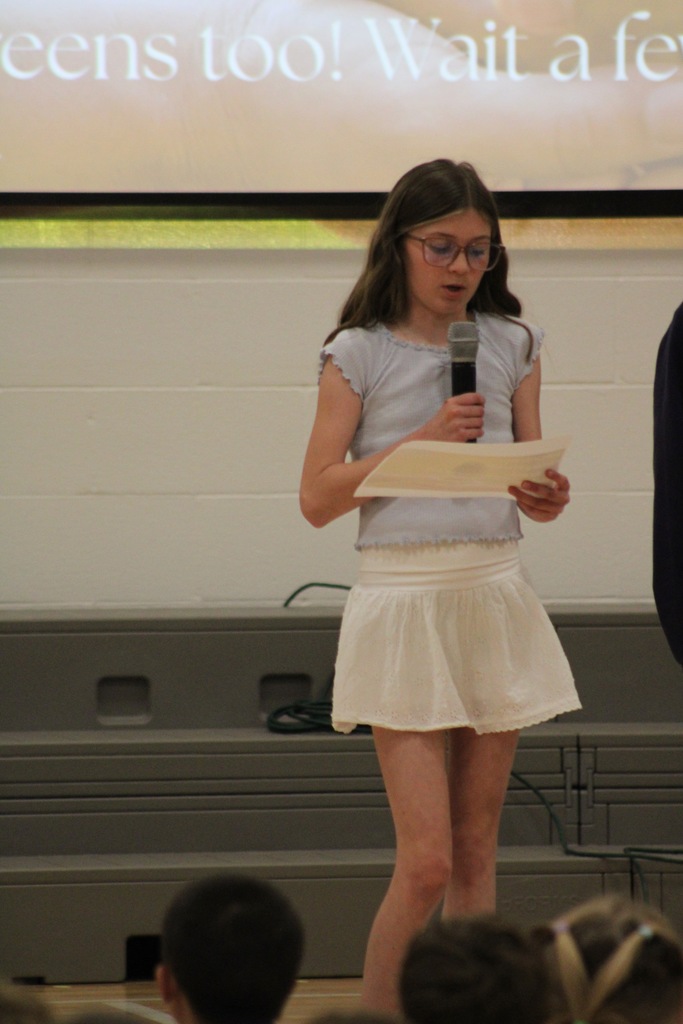 A girl in a light blue shirt stands at the front holding a microphone reading her poem.