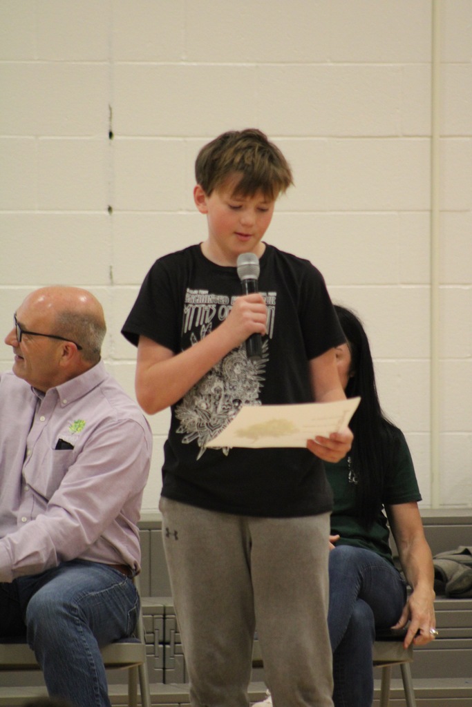 A boy in a black shirts stands at the front with a microphone reading his poem.