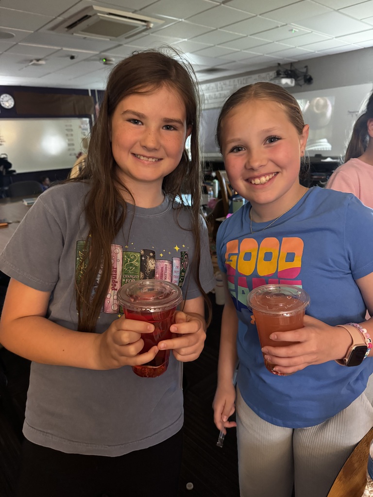 Two smiling young girls in a classroom holding clear plastic cups filled with red and orange beverages.