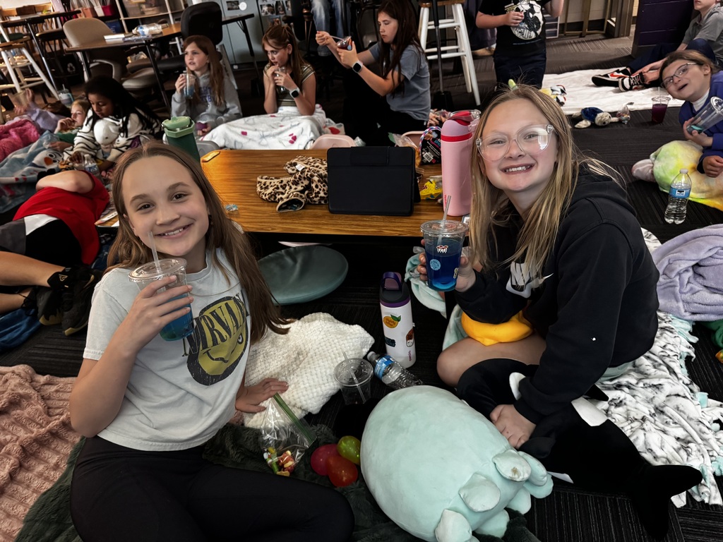 Two girls in the foreground sitting on the floor with blue drinks and a large plush toy, while other students relax in the background.
