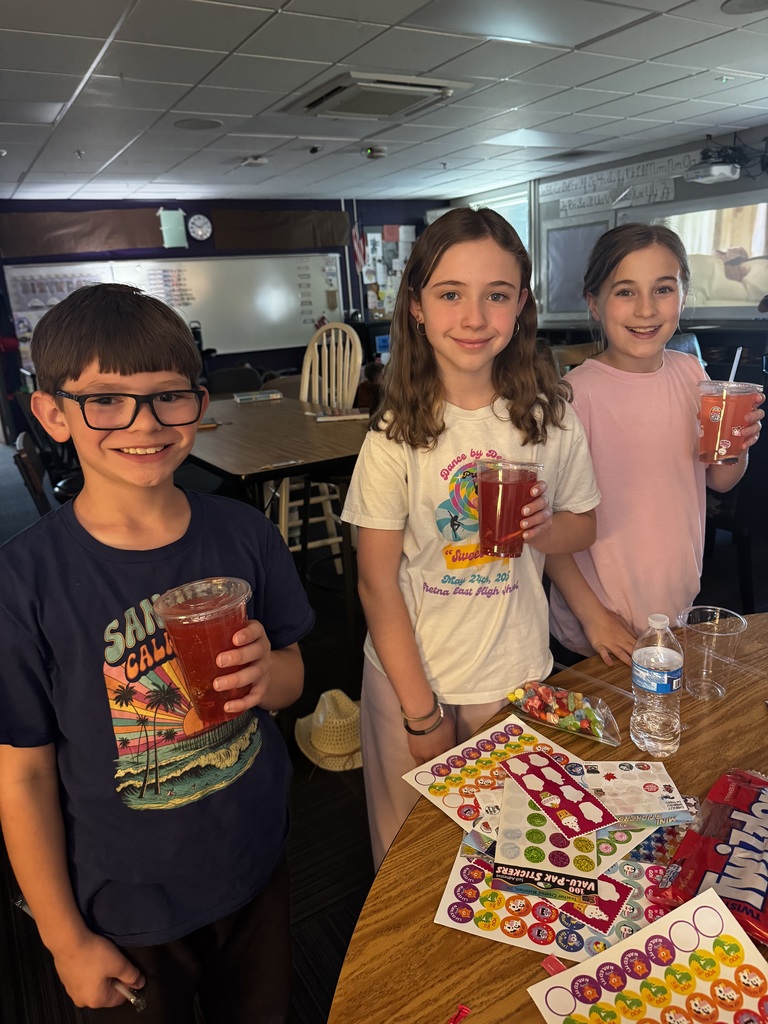 Three children standing behind a table scattered with stickers and candy, each holding a cup of red juice.