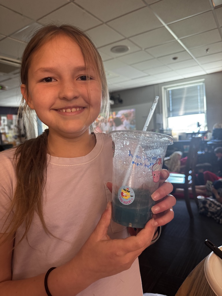 A close-up of a smiling girl holding a clear plastic cup with a blue drink and decorative stickers.