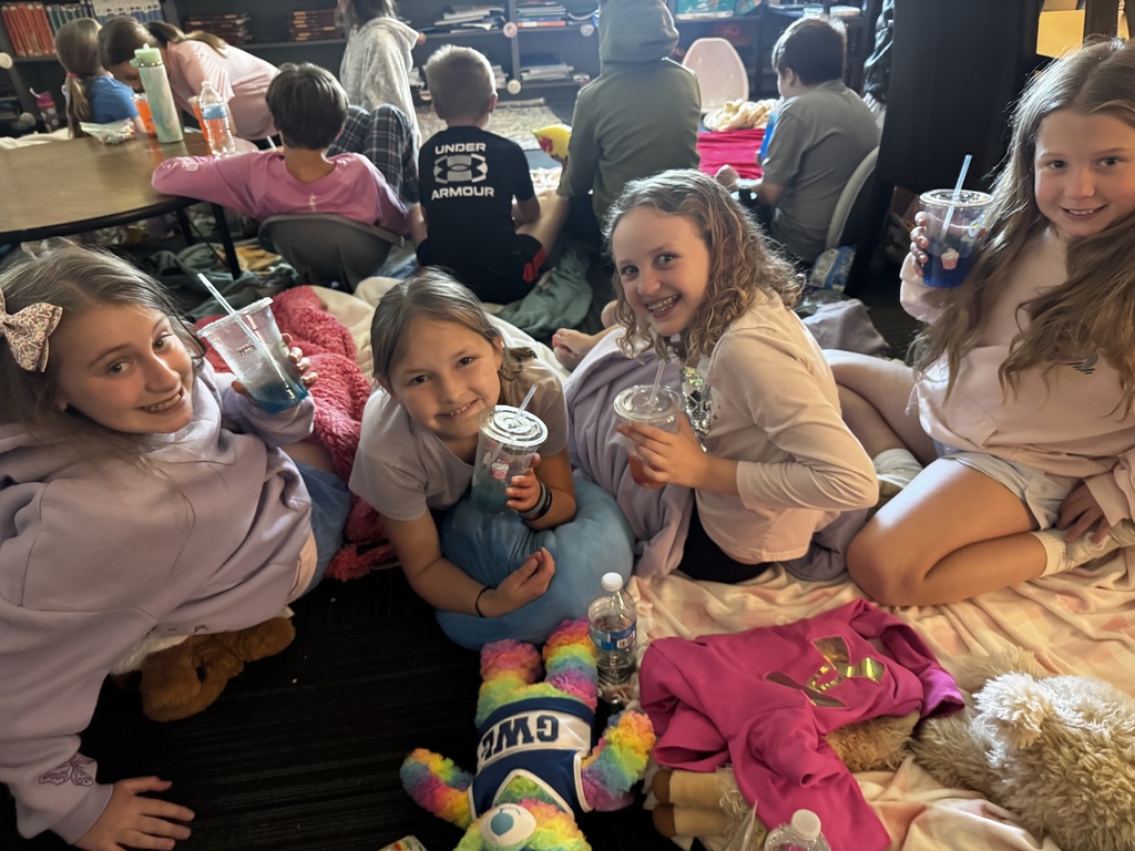 A group of children sitting on blankets on a classroom floor with pillows, stuffed animals, and colorful drinks.