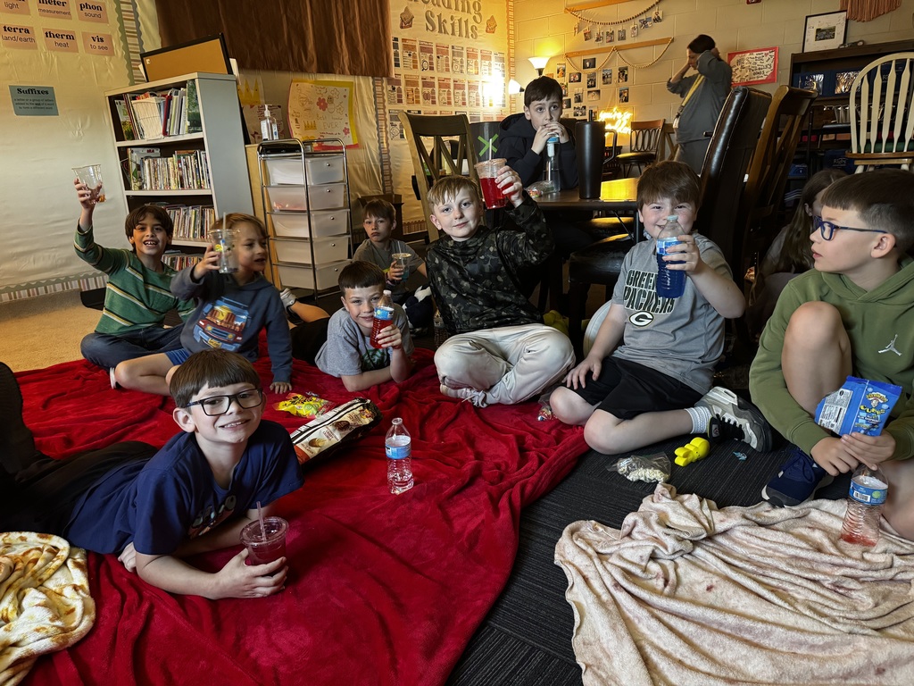 Several boys sitting on a large red blanket on the floor, holding up their colorful drinks and water bottles.