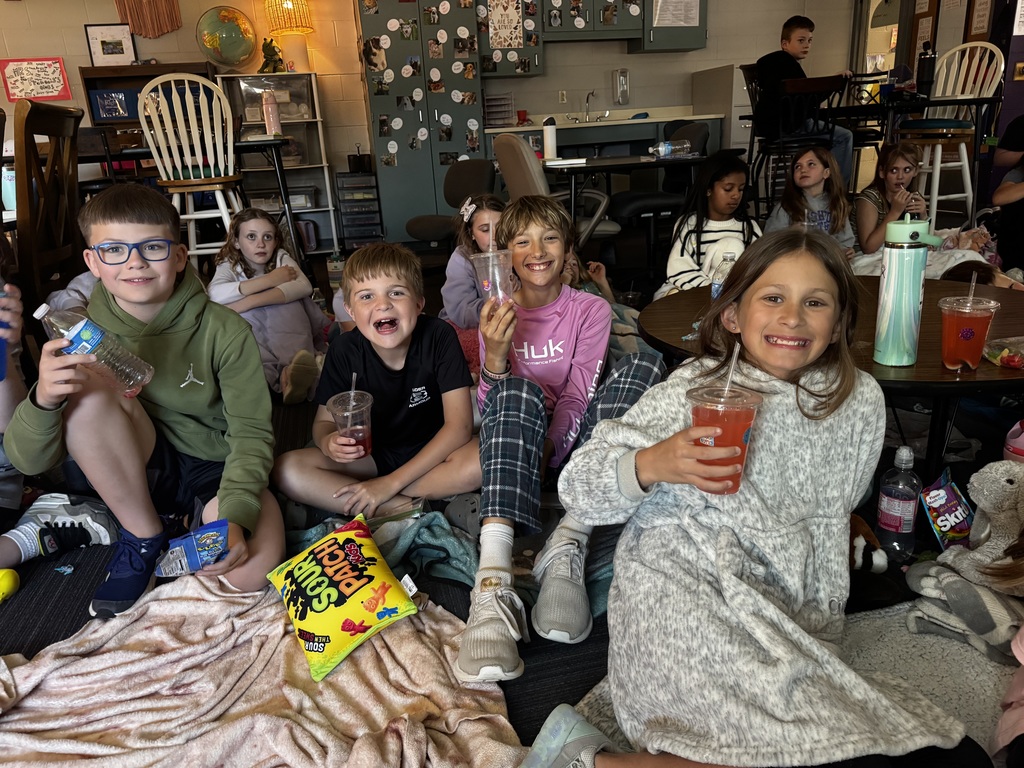 A group of students sitting on the floor amongst snacks and blankets, smiling and holding up their red and purple drinks.