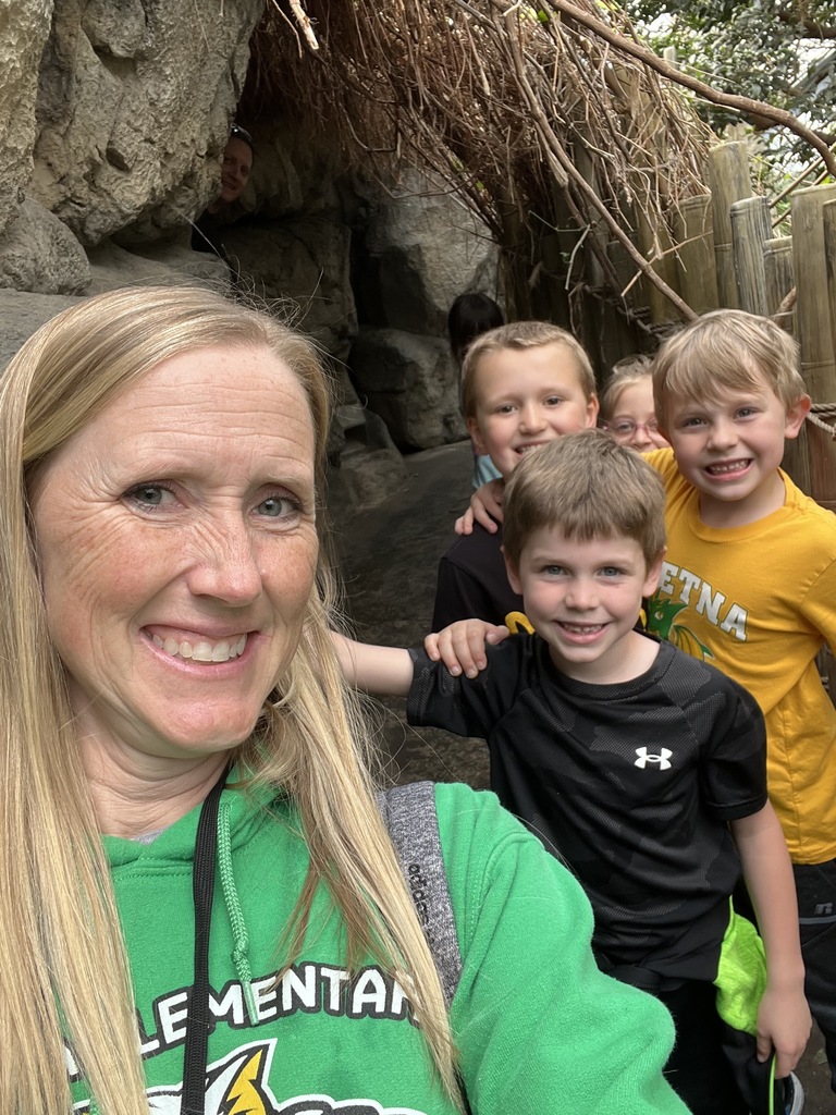A woman in a green hoodie takes a smiling selfie with four excited children in a rocky, tunnel-like outdoor walkway.
