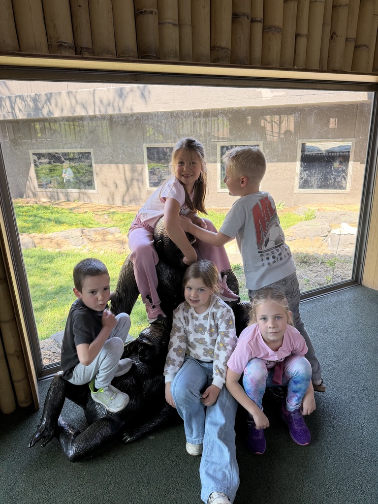 Five children play on and around a black gorilla statue inside a zoo building with large windows overlooking a grassy habitat.