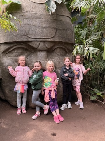 Five young girls line up and pose in front of the same large stone head sculpture seen in the jungle exhibit.