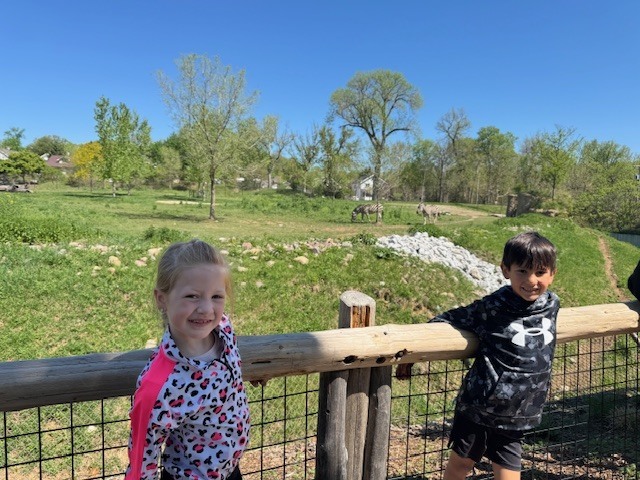 A young girl and boy lean against a wooden fence at a zoo, with zebras grazing in a grassy field in the background.