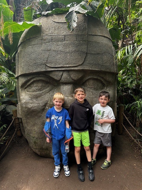 Three young boys pose in front of a massive, ancient-style stone head sculpture surrounded by jungle foliage.