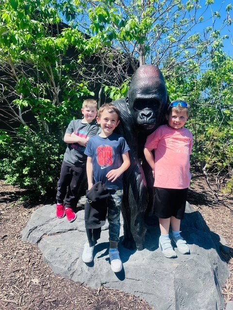 Three young boys smile while posing with a life-sized black gorilla statue outdoors under a bright blue sky.