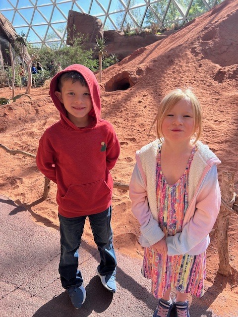 Two children stand on a sandy path inside a desert-themed dome exhibit with reddish rock formations.