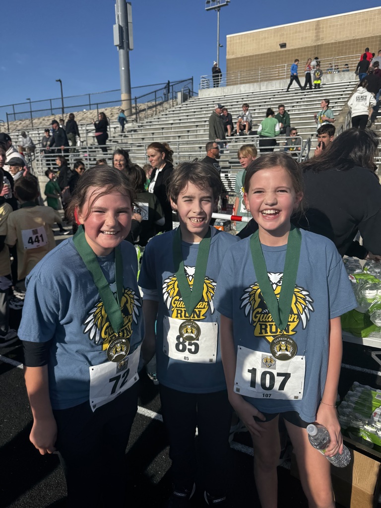 Three smiling young children pose for a photo outdoors in the bright sun. They are wearing blue race shirts, green-ribboned medals, and white race bibs with numbers 72, 85, and 107. A concrete stadium is in the background.