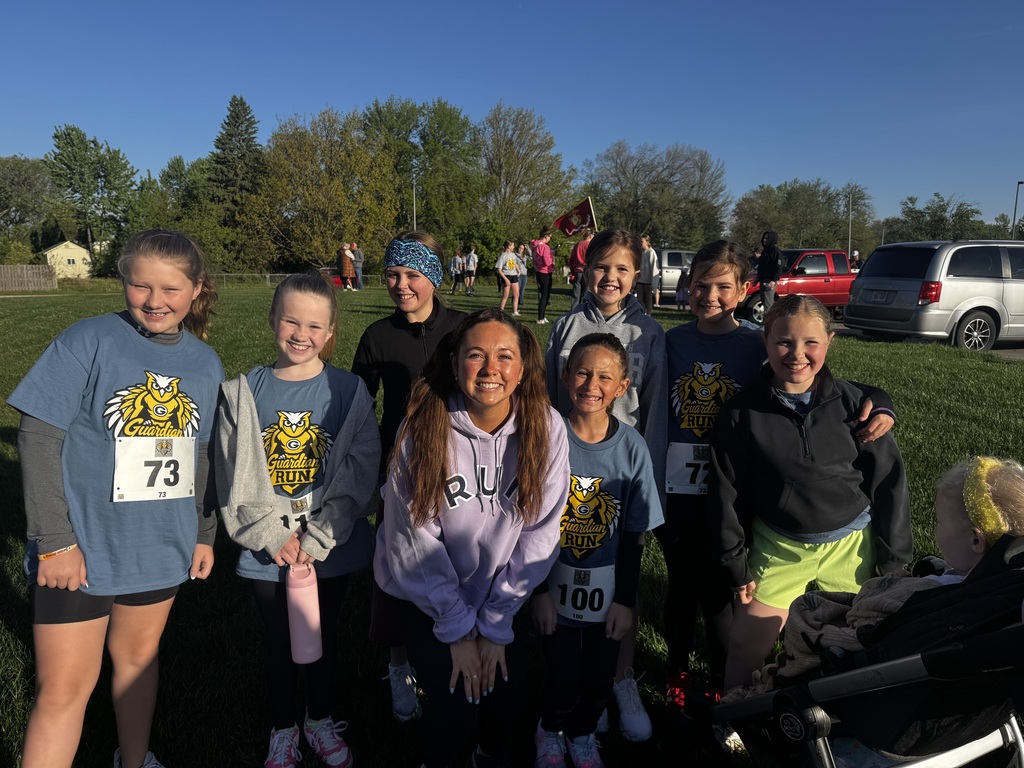 Seven smiling girls and one woman in a lavender hoodie gather closely on a sunny lawn. Most wear blue owl-logo shirts and race bibs (like 73, 72, 100). The woman is crouched, and a small child in a stroller is on the far right.