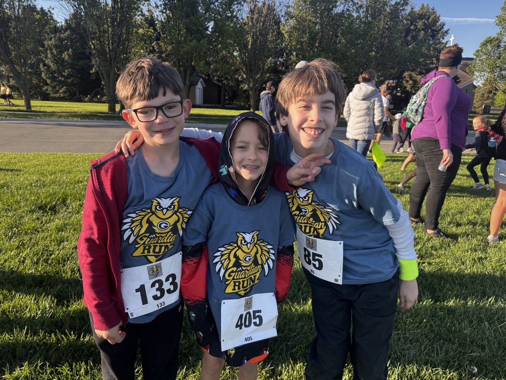 Three young boys pose together on a green lawn, their blue shirts featuring an owl logo. Two wear glasses, one wears a hoodie, and their white bibs display numbers 133, 405, and 85. A busy road is behind them.