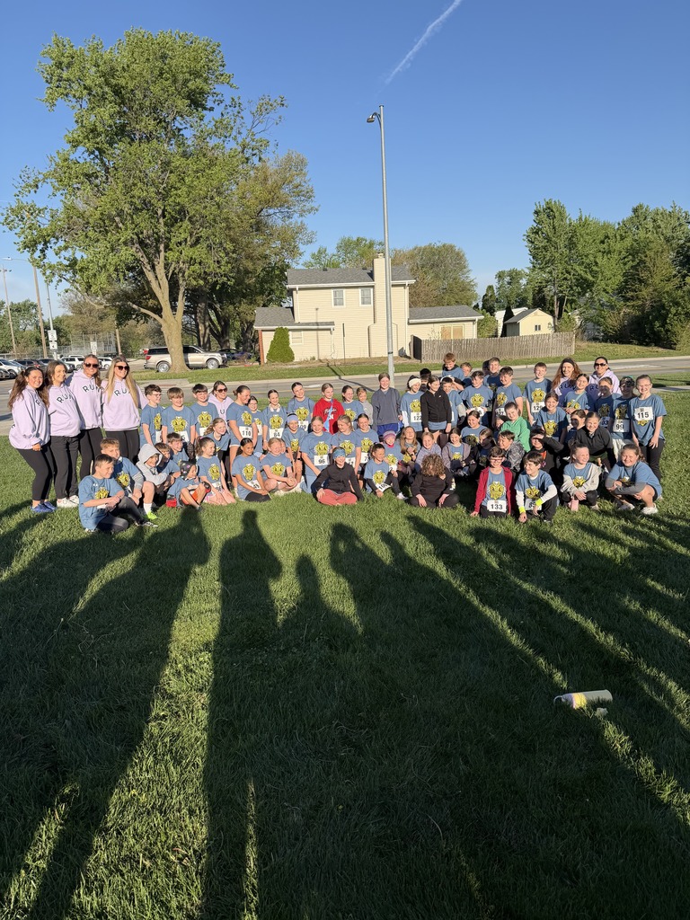 A wide shot of a large group of children and several adults in lavender hoodies gathered on a grassy field in front of a house. The bright sun casts long, dark shadows toward the viewer. Most children wear blue shirts, and a road is behind them.