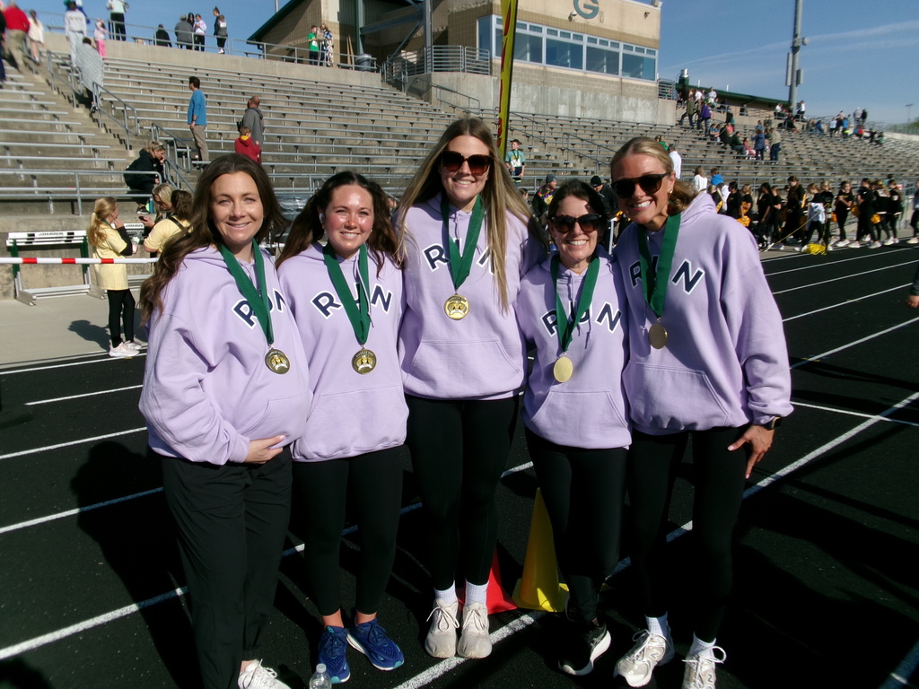 Five women stand on an outdoor black running track, all wearing matching lavender hoodies, black pants, and large gold medals with green ribbons. They are in front of concrete stadium bleachers under a clear blue sky.
