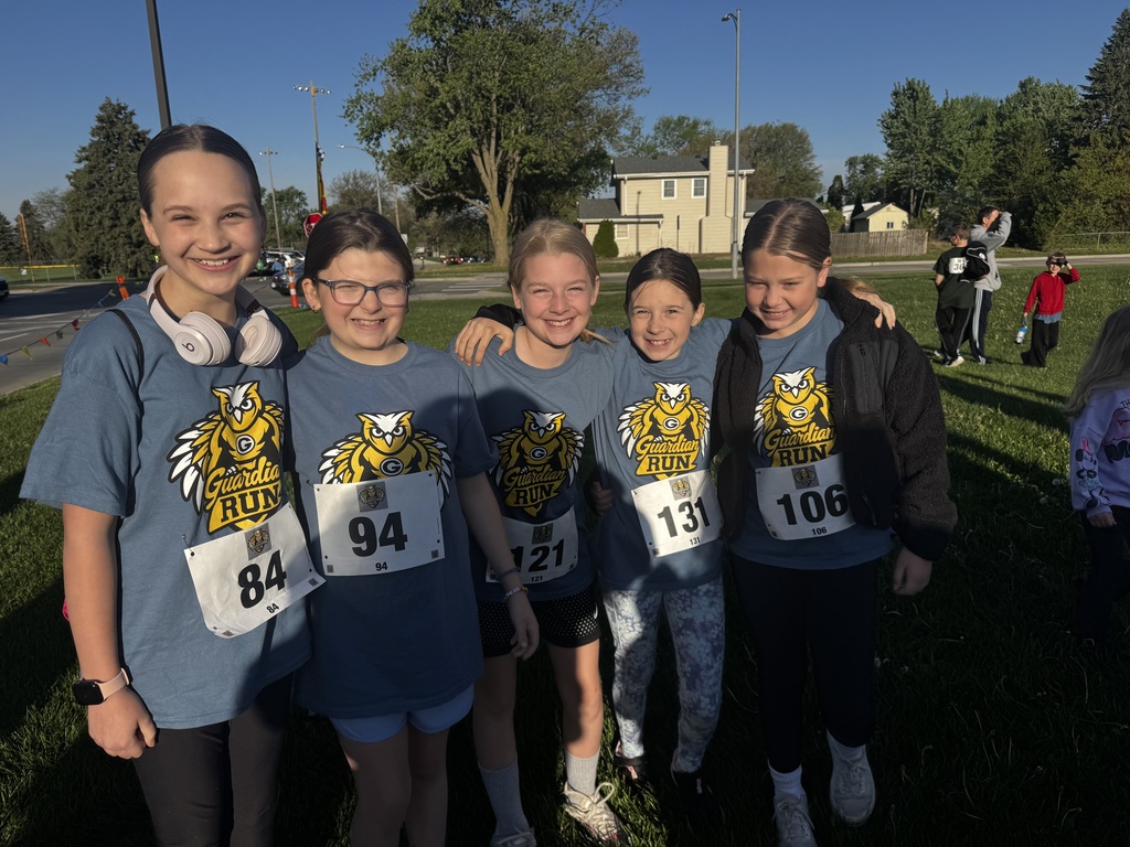 Five young girls pose together on a grassy area next to a road, all wearing blue race shirts with an owl logo and white bibs (numbers 84, 94, 121, 131, 106). The bright sun is low, making them smile. Trees and a house are in the distance.