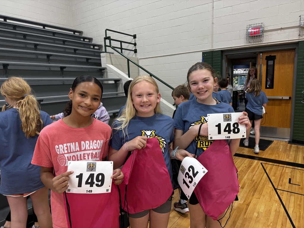 Inside a gymnasium with bleachers, three young girls hold up their white race bibs (numbers 149, 132, 148) and bright pink drawstring bags. A wood floor is visible, and others stand in the background.