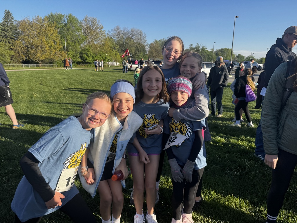 Six young girls, with matching blue owl-logo shirts and white race bibs (numbers 13, 92), hug tightly together on a grassy field. Two wear winter hats. Bright sunlight casts long shadows, and other people are in the background.