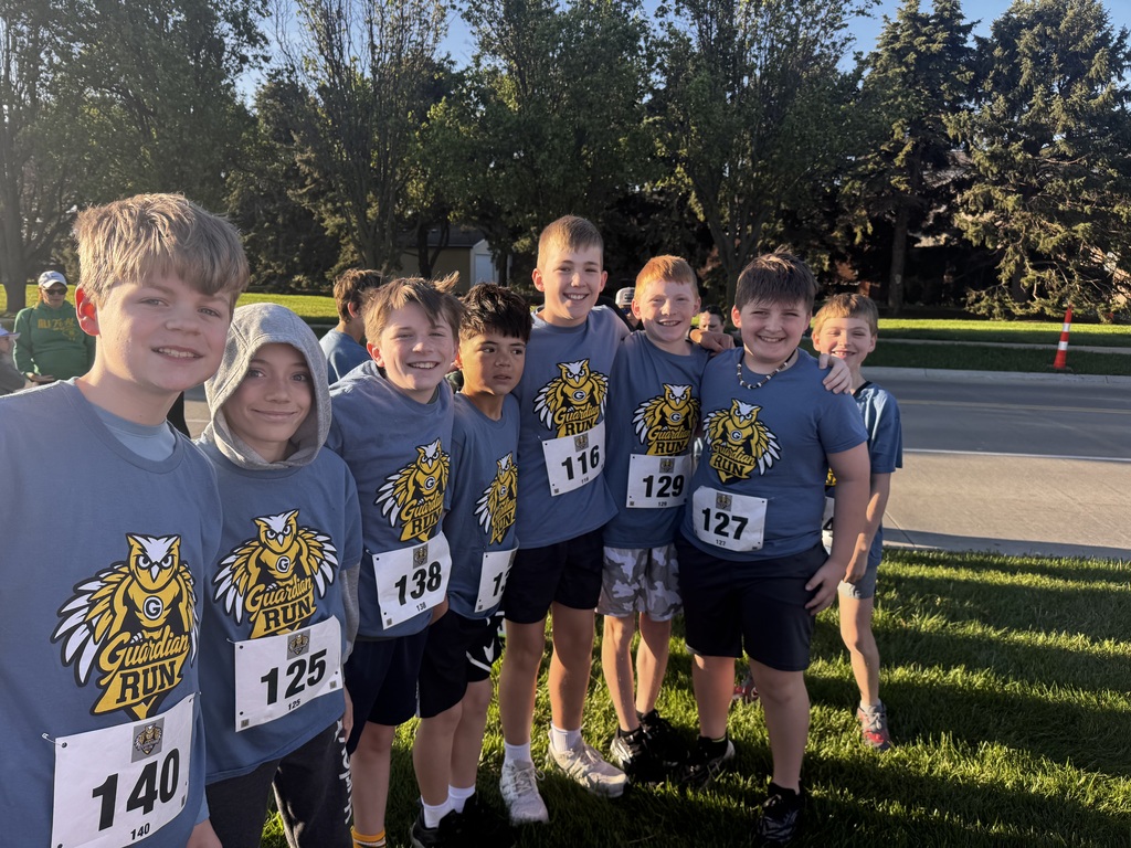 Seven young boys, arm-in-arm, stand on a green lawn next to a street. All wear blue race shirts featuring a large owl logo and white bibs (numbers 140, 125, 138, 116, 129, 127). The bright sun illuminates them.